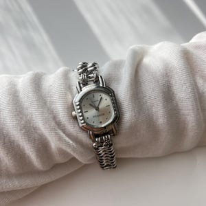 May include: A silver-toned wristwatch with a rectangular face and a linked band. The watch face is white with black hands and hour markers. The band has a textured, woven appearance.