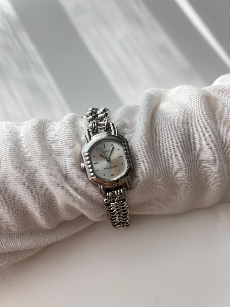 May include: A silver-toned wristwatch with a rectangular face and a linked band. The watch face is white with black hands and hour markers. The band has a textured, woven appearance.