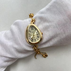 Pode incluir: Um rel&oacute;gio de pulso dourado com um design &uacute;nico de mostrador de rel&oacute;gio derretido. O rel&oacute;gio apresenta um mostrador creme com algarismos romanos e ponteiros pretos. A pulseira do rel&oacute;gio tamb&eacute;m &eacute; dourada, com um detalhe de corrente decorativa. O rel&oacute;gio est&aacute; rotulado como "BARSH QUARTZ".
