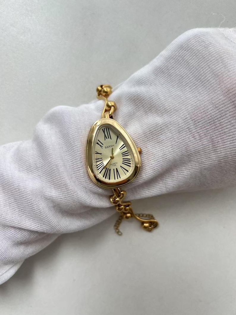 Pode incluir: Um rel&oacute;gio de pulso dourado com um design &uacute;nico de mostrador de rel&oacute;gio derretido. O rel&oacute;gio apresenta um mostrador creme com algarismos romanos e ponteiros pretos. A pulseira do rel&oacute;gio tamb&eacute;m &eacute; dourada, com um detalhe de corrente decorativa. O rel&oacute;gio est&aacute; rotulado como "BARSH QUARTZ".