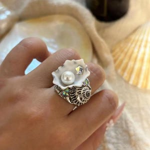 Könnte beinhalten: Ein silberner Ring mit einem weißen Muschel-Design. Der Ring hat eine Perle und einen Strassstein in der Mitte der Muschel.