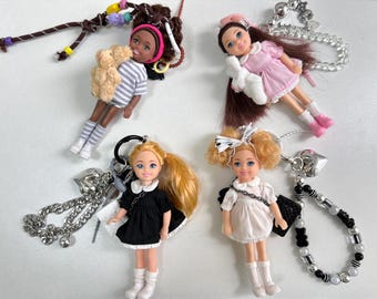 Könnte beinhalten: Vier kleine Puppen-Schlüsselanhänger mit verschiedenen Outfits und Accessoires. Die Puppen tragen Kleider und haben langes Haar. Die Schlüsselanhänger haben verschiedene Anhänger, darunter Herzen, Ketten und Perlen.
