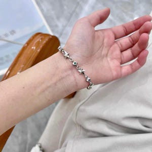 Może przedstawiać: Srebrna bransoletka z zawieszkami w kształcie gwiazd i małymi kamieniami szlachetnymi. Bransoletka jest noszona na nadgarstku, podkreślając jej delikatny design i odblaskowe właściwości. Ogniwa bransoletki są widoczne, co dodaje jej skomplikowanego wyglądu.
