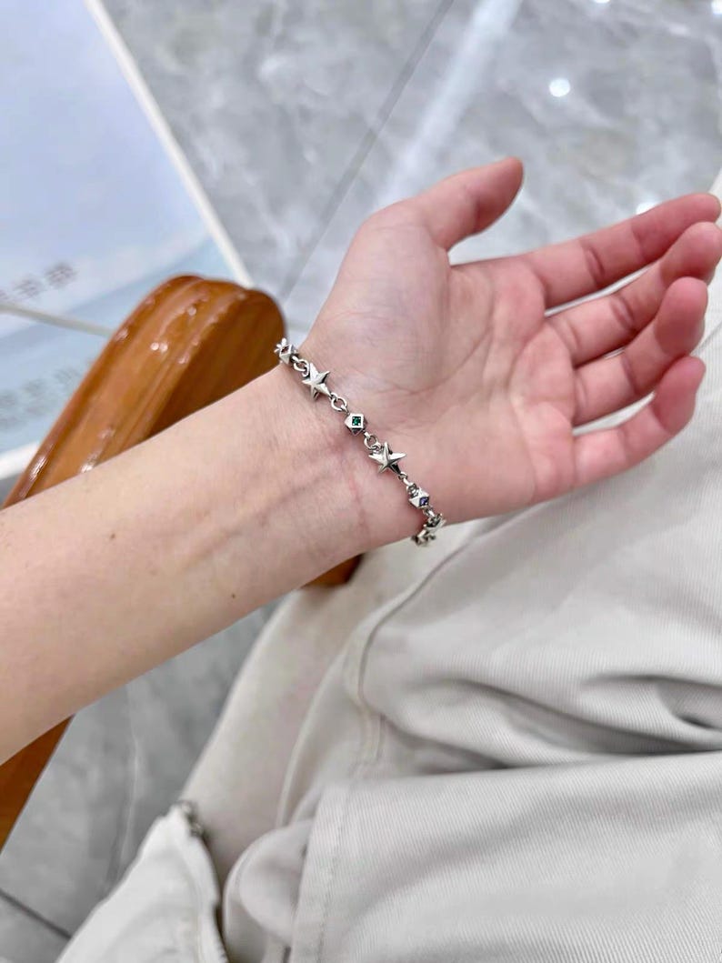 Może przedstawiać: Srebrna bransoletka z zawieszkami w kształcie gwiazd i małymi kamieniami szlachetnymi. Bransoletka jest noszona na nadgarstku, podkreślając jej delikatny design i odblaskowe właściwości. Ogniwa bransoletki są widoczne, co dodaje jej skomplikowanego wyglądu.