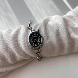 May include: Silver-toned wristwatch with an oval black face and Roman numeral hour markers. The watch band is a delicate chain design. The watch face has the word "NAIDU" printed on it.