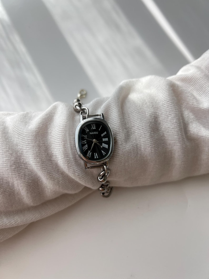 May include: Silver-toned wristwatch with an oval black face and Roman numeral hour markers. The watch band is a delicate chain design. The watch face has the word "NAIDU" printed on it.