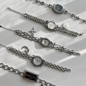 May include: Five silver-toned wristwatches with various face shapes and chain styles. Each watch features a different face color, including black, white, and cream. Some watches have charms like a heart, arrow, moon, and cross. The watches are displayed on a white surface.