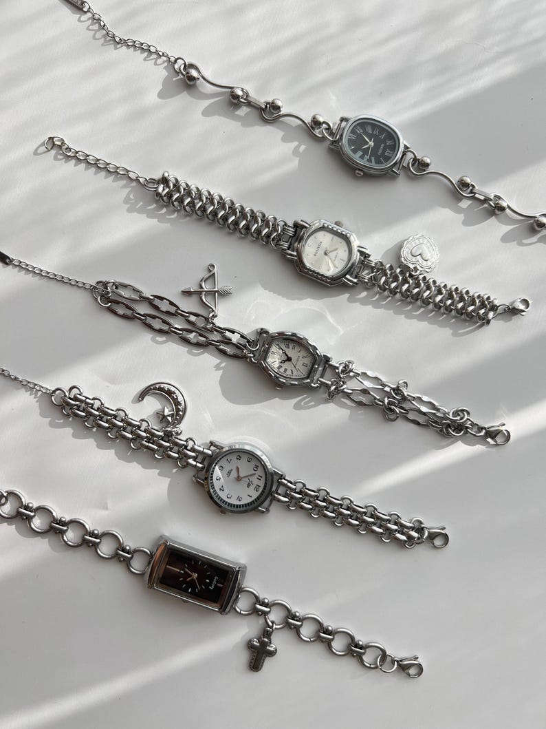 May include: Five silver-toned wristwatches with various face shapes and chain styles. Each watch features a different face color, including black, white, and cream. Some watches have charms like a heart, arrow, moon, and cross. The watches are displayed on a white surface.