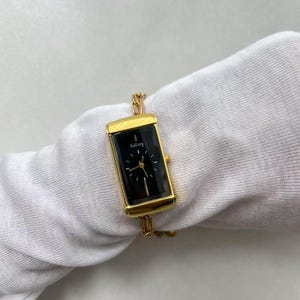 Pode incluir: Rel&oacute;gio retangular dourado com mostrador preto e ponteiros dourados. O rel&oacute;gio tem o nome da marca "GoGoey" impresso no mostrador. O rel&oacute;gio &eacute; preso a uma pulseira de corrente dourada.