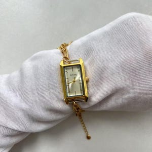 Pode incluir: Rel&oacute;gio de pulso retangular dourado com mostrador claro e pulseira de elos. O mostrador do rel&oacute;gio tem o nome da marca "GoGory" impresso. O rel&oacute;gio &eacute; exibido em uma superf&iacute;cie branca.