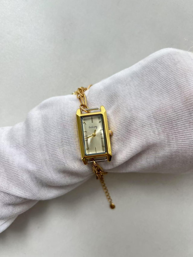 Pode incluir: Rel&oacute;gio de pulso retangular dourado com mostrador claro e pulseira de elos. O mostrador do rel&oacute;gio tem o nome da marca "GoGory" impresso. O rel&oacute;gio &eacute; exibido em uma superf&iacute;cie branca.