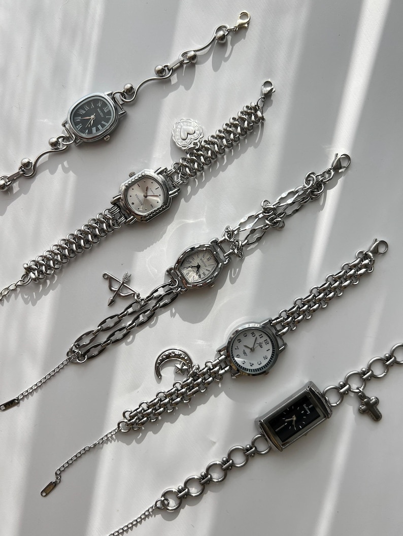 May include: A collection of silver-toned wristwatches with various chain designs. Each watch features a different face shape and style, including oval, rectangular, and round. Some watches have decorative charms like a heart, crescent moon, and cross.
