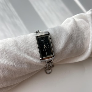 May include: A rectangular silver watch with a black face and the word "GoGoey" printed on the top. The watch has a silver chain band and is displayed on a white fabric.