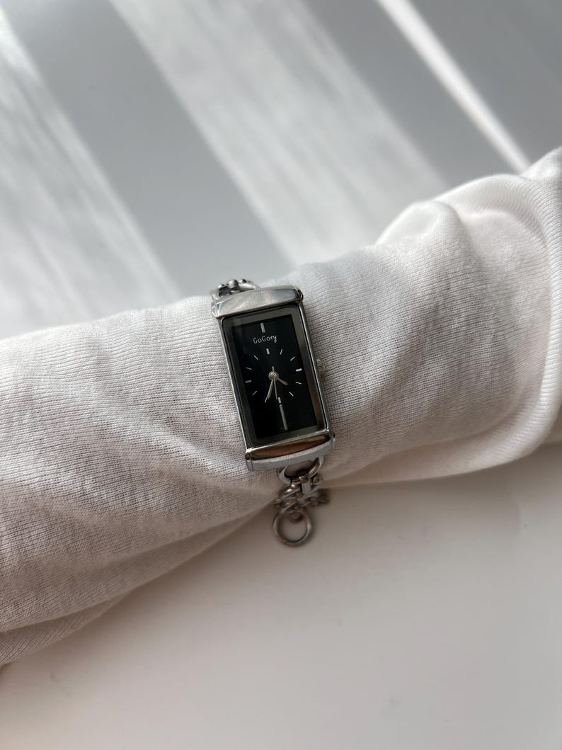 May include: A rectangular silver watch with a black face and the word "GoGoey" printed on the top. The watch has a silver chain band and is displayed on a white fabric.