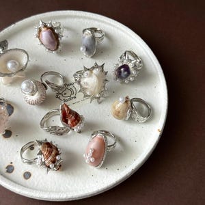 Puede incluir: Una colección de anillos de plata con diversas conchas naturales y pequeñas perlas blancas. Los anillos están dispuestos sobre un plato circular blanco moteado que incluye una pequeña tarjeta con el texto Nola Toams y un dibujo de un pez.
