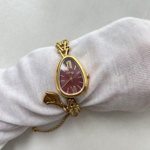 Pode incluir: Um rel&oacute;gio de pulso dourado com mostrador bord&ocirc; e ponteiros dourados. O rel&oacute;gio tem um formato &uacute;nico de gota e uma delicada pulseira de corrente. O mostrador exibe o algarismo romano XII. O rel&oacute;gio est&aacute; sobre um tecido branco.