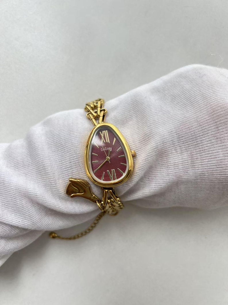 Pode incluir: Um rel&oacute;gio de pulso dourado com mostrador bord&ocirc; e ponteiros dourados. O rel&oacute;gio tem um formato &uacute;nico de gota e uma delicada pulseira de corrente. O mostrador exibe o algarismo romano XII. O rel&oacute;gio est&aacute; sobre um tecido branco.