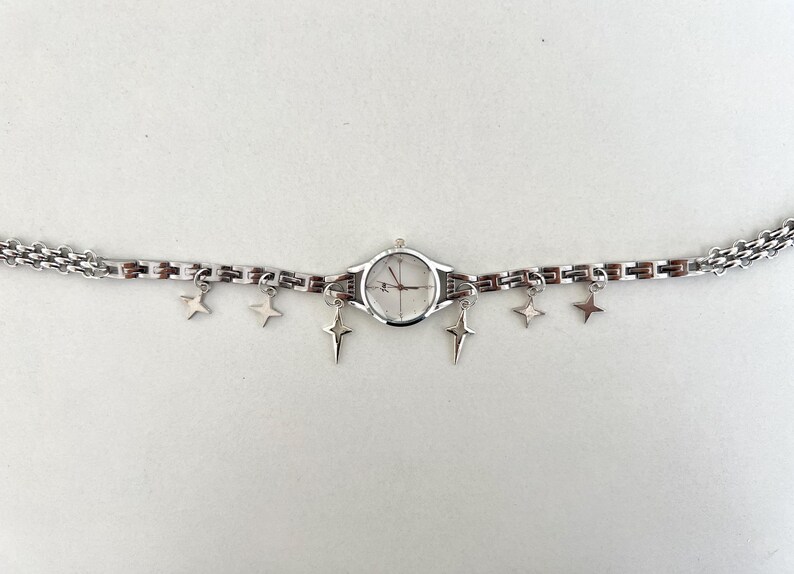 Op de afbeelding: Een zilveren horloge met een witte wijzerplaat en een zilveren kettingarmband met stervormige bedels.