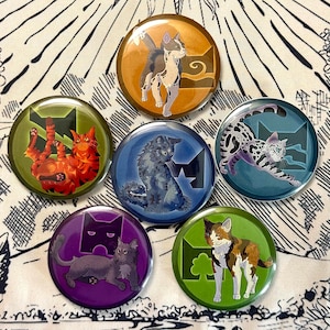 May include: A collection of seven round pinback buttons featuring cat illustrations in various poses and colors. Each button has a different cat design, with a logo and background color. The buttons are arranged on a patterned white surface.