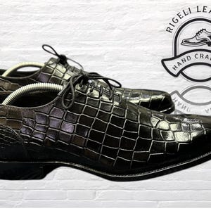 May include: Black leather dress shoes with a crocodile pattern. The shoes have a lace-up closure and a pointed toe. The shoes are made by Rigelli Leather and are hand-crafted.