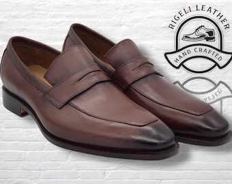 Könnte beinhalten: Ein Paar braune Leder-Penny-Loafer mit dunkler Spitze. Die Schuhe haben ein klassisches Design mit einem Riemen über der Oberseite. Das Bild enthält ein Logo mit der Aufschrift "RIGELI LEATHER HAND CRAFTED".