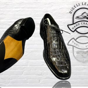 May include: A pair of black leather dress shoes with a crocodile pattern. The shoes are hand-crafted and feature a leather sole. The image includes the text "Rigelli Leather Hand Crafted".