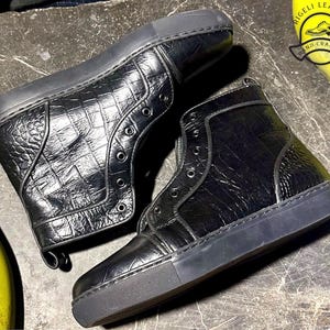 May include: Black high-top sneakers with a textured, crocodile-like pattern. The sneakers have a rubber sole and eyelets for laces. The image includes a logo that says "RIGELI LEATHER HAND CRAFTED".