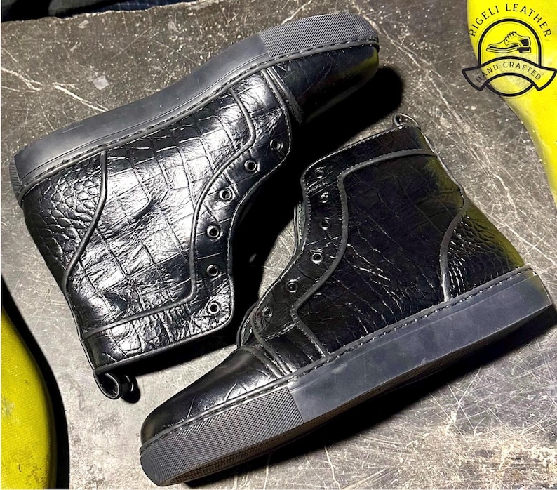 May include: Black high-top sneakers with a textured, crocodile-like pattern. The sneakers have a rubber sole and eyelets for laces. The image includes a logo that says "RIGELI LEATHER HAND CRAFTED".