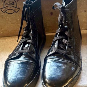 May include: A pair of black leather boots with a textured, crocodile-like pattern. The boots have black laces and soles. The brand logo "Rigeli Leather, Hand Crafted" is visible in the upper left corner.