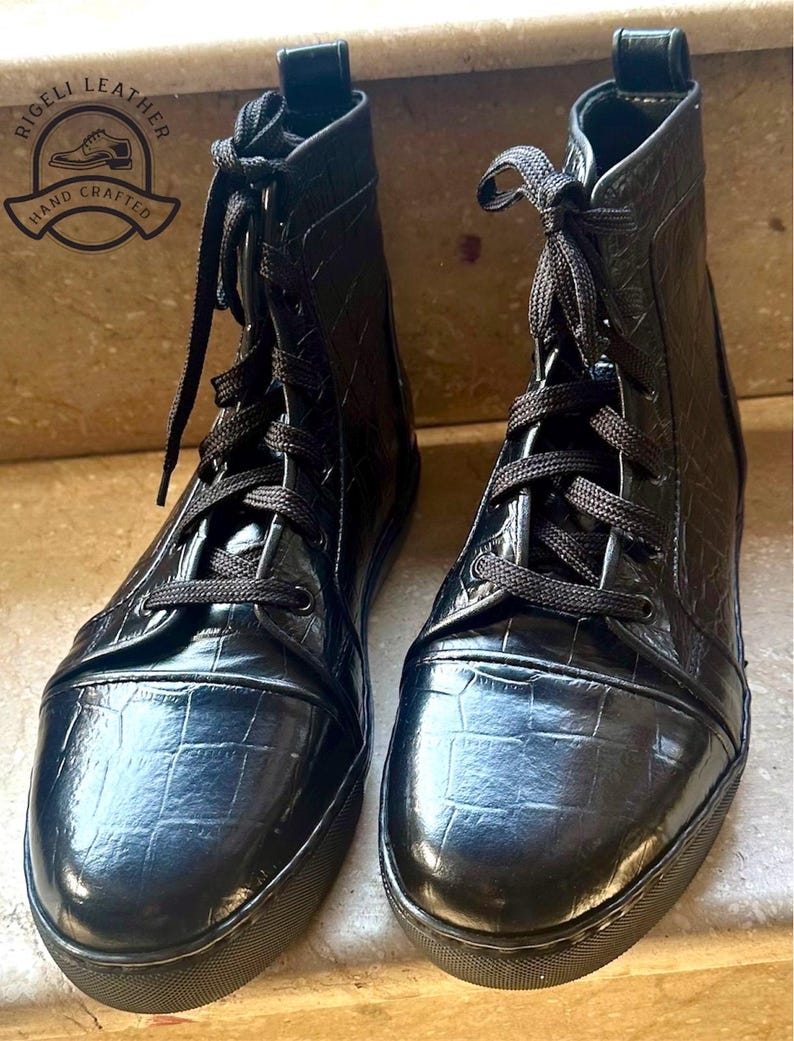May include: A pair of black leather boots with a textured, crocodile-like pattern. The boots have black laces and soles. The brand logo "Rigeli Leather, Hand Crafted" is visible in the upper left corner.