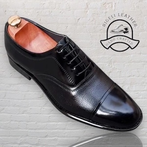 May include: Black leather lace-up Oxford shoes with a cap toe. The shoes have a textured leather upper and a polished finish. The brand name "RIGELLI LEATHER HAND CRAFTED" is printed on a circular label.
