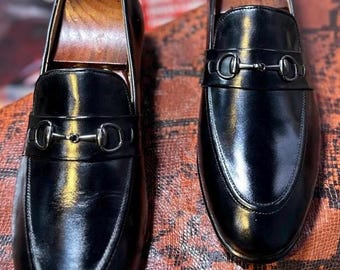 Könnte beinhalten: Ein Paar schwarze Leder-Loafer mit polierter Oberfläche, abgerundeter Zehenpartie und einem silberfarbenen Metall-Trensendetail über dem Vorderblatt. Jeder Schuh enthält einen hölzernen Schuhspanner, um die Form des Schuhs zu bewahren.