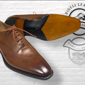 May include: A pair of brown leather dress shoes with intricate brogue detailing and matching laces. One shoe is upright, while the other is upside down, showcasing the sole. The shoes are set against a white brick wall.