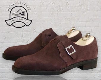 Könnte beinhalten: Ein Paar braune Wildlederschuhe mit Monk-Riemen und einer silbernen Schnalle. Die Schuhe sind handgefertigt und haben eine Ledersohle. Die Schuhe stehen auf einem weißen Ziegelhintergrund. Der Text "RIGELLI LEATHER HAND CRAFTED" befindet sich in einem Kreis über den Schuhen.
