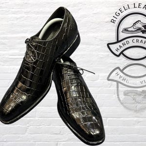May include: A pair of black leather dress shoes with a crocodile pattern. The shoes have lace-up closures and are made by Rigelli Leather, a company that specializes in hand-crafted footwear.