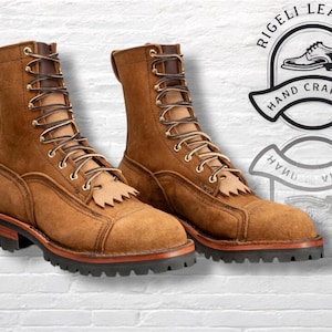 May include: A pair of brown leather work boots with a lace-up design and a rugged sole. The boots have a mid-calf height and a decorative fringe on the tongue. The boots are labeled "Rigeli Leather, Hand Crafted."