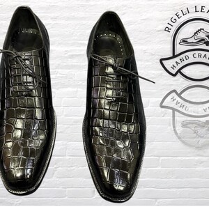 May include: A pair of black leather dress shoes with a crocodile pattern. The shoes have lace-up closures and are hand-crafted. The image also includes a logo that says "Rigelli Leather Hand Crafted".