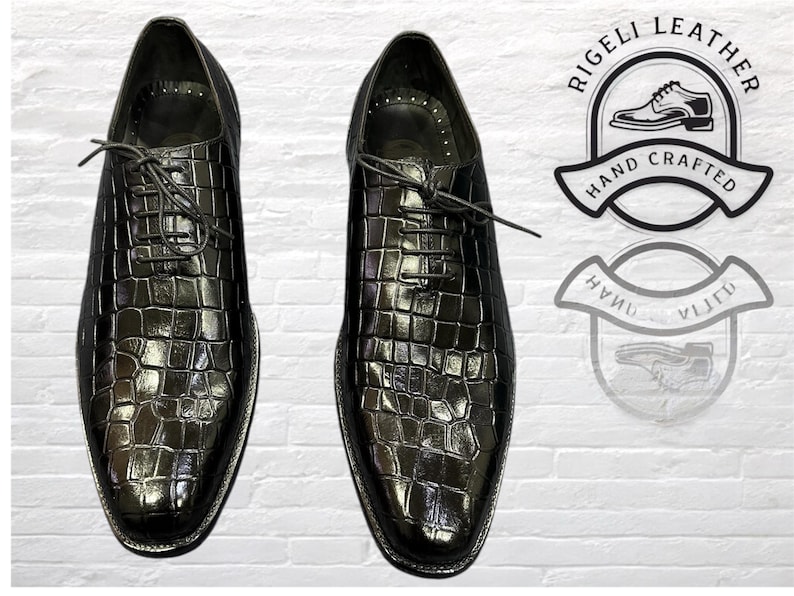 May include: A pair of black leather dress shoes with a crocodile pattern. The shoes have lace-up closures and are hand-crafted. The image also includes a logo that says "Rigelli Leather Hand Crafted".