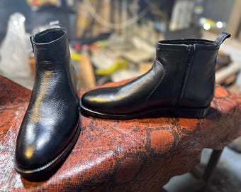 Könnte beinhalten: Ein Paar schwarze Lederstiefeletten mit glatter, polierter Oberfläche, abgerundeter Zehenpartie, seitlichem Reißverschluss und einer kleinen Zugschlaufe an der Ferse. Die Stiefel haben einen niedrigen Absatz und eine strukturierte Form mit sichtbaren Nähten.