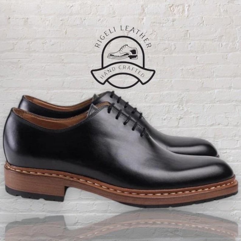 May include: Black leather lace-up shoes with a brown leather sole. The shoes have a stitched detail around the sole. The shoes are labeled "Rigelli Leather Hand Crafted".