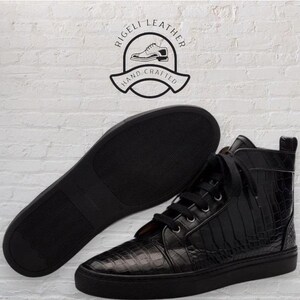 May include: Black high-top sneakers with a textured, crocodile-like pattern. The sneakers have black laces and soles. The sole of a matching sneaker is visible next to the shoe. The image includes a logo that says "Rigeli Leather Hand Crafted".