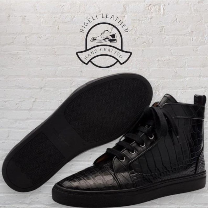 May include: Black high-top sneakers with a textured, crocodile-like pattern. The sneakers have black laces and soles. The sole of a matching sneaker is visible next to the shoe. The image includes a logo that says "Rigeli Leather Hand Crafted".