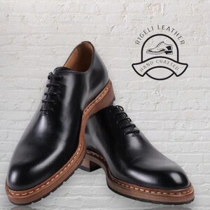 May include: Two black leather lace-up shoes with brown leather soles. The shoes have a stitched edge around the sole. The shoes are on a white brick background. The text "Rigelli Leather Hand Crafted" is in a circle above the shoes.