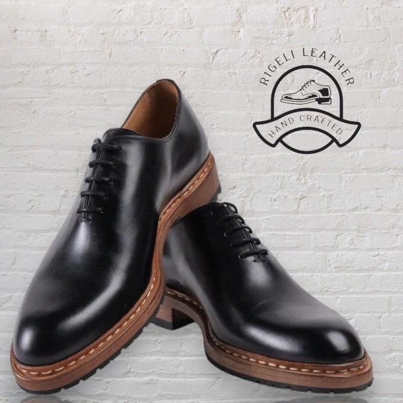 May include: Two black leather lace-up shoes with brown leather soles. The shoes have a stitched edge around the sole. The shoes are on a white brick background. The text "Rigelli Leather Hand Crafted" is in a circle above the shoes.