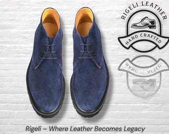 Könnte beinhalten: Ein Paar marineblaue Wildleder-Chukka-Stiefel mit schwarzen Sohlen und Schnürsenkeln. Der Innenraum ist mit gelbem Leder gefüttert. Das Bild enthält den Text "Rigeli - Where Leather Becomes Legacy" und ein Logo "Hand Crafted".