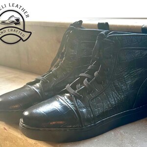May include: Pair of black leather high-top sneakers with a textured, crocodile-like pattern. The sneakers have black laces and soles. The image includes a logo that says "Rigeli Leather" and "Hand Crafted".