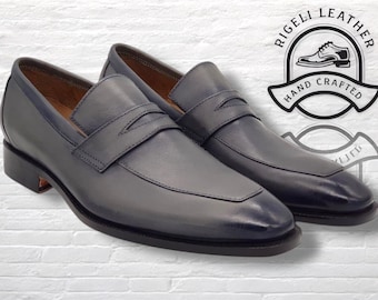 Könnte beinhalten: Ein Paar blau-graue Leder-Penny-Loafer. Die Schuhe haben ein klassisches Design mit einem Riemen über dem Spann und einem niedrigen Absatz. Das Bild enthält ein Logo mit der Aufschrift "RIGELI LEATHER HAND CRAFTED".