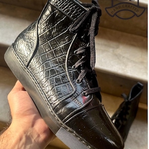 Op de afbeelding: Zwarte leren high-top sneakers met een textuurpatroon dat op krokodillenleer lijkt. De sneakers hebben zwarte veters en een zwarte zool. De afbeelding bevat een logo met de tekst "RIGELI LEATHER HAND CRAFTED".