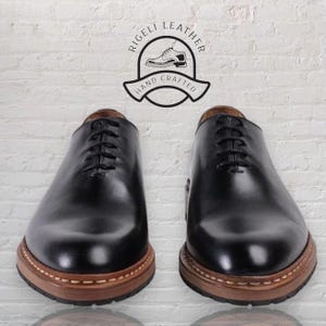 May include: A pair of black leather lace-up shoes with brown soles. The shoes have a stitched detail around the sole and are made by Rigelli Leather, Hand Crafted.