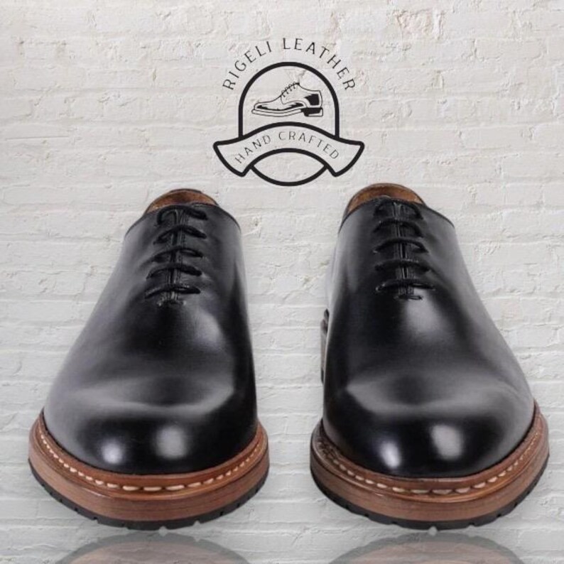 May include: A pair of black leather lace-up shoes with brown soles. The shoes have a stitched detail around the sole and are made by Rigelli Leather, Hand Crafted.